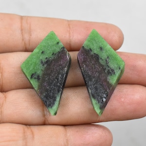Pode incluir: Duas pedras preciosas em forma de diamante. Cada pedra apresenta uma metade superior verde vibrante e uma base escura e manchada de preto e roxo. As pedras são seguradas em uma mão, mostrando sua aparência natural e não polida. As pedras preciosas são provavelmente para fazer joias.