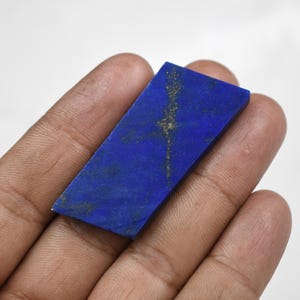 May include: A rectangular, polished lapis lazuli gemstone. The stone is a deep, vibrant blue with streaks of gold and lighter blue veining. The gemstone is held in a hand against a white background.