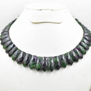 May include: A faceted gemstone necklace displayed on a white mannequin. The necklace features a series of dark green and purple stones. The stones are cut into a geometric shape, creating a unique and eye-catching design. The necklace is a statement piece.