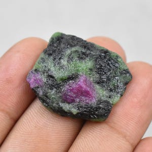 Puede incluir: Una gema en bruto con un vibrante centro magenta, rodeada de minerales verdes y negros. La piedra tiene una superficie texturizada y una forma irregular, mostrando variaciones naturales de color y patrón. Este espécimen de rubí en bruto es único.