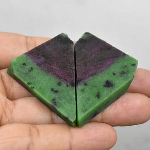 Puede incluir: Dos piezas triangulares de piedra preciosa zoisita con superficie pulida. Cada pieza presenta un color base verde vibrante con un centro púrpura intenso e inclusiones negras dispersas, dispuestas lado a lado formando una figura de corazón.