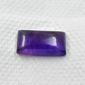 May include: A rectangular, polished amethyst gemstone with rounded corners. The gem displays a deep purple hue with a glossy surface, reflecting light. The stone is set against a white, textured background.