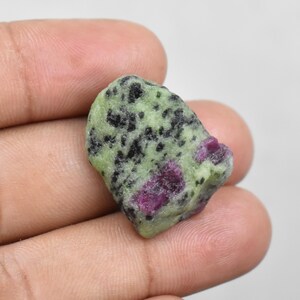 Peut inclure: Une pierre précieuse brute avec une surface verte et noire mouchetée, parsemée d'inclusions rouges vives. La pierre est tenue dans une main, mettant en valeur sa texture naturelle et non polie et ses variations de couleur. Spécimen minéral unique.