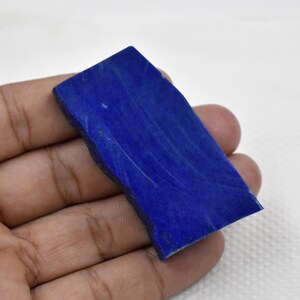 以下が含まれることがあります： 長方形の濃い青色の宝石で、粗く不均一なエッジがあります。石は研磨された表面で、わずかに白い静脈があります。宝石は白い背景に対して手で持たれています。