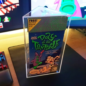 May include: A clear plastic display case with a green, black, and white video game box inside. The box features the title "Day of the Tentacle" and a cartoon illustration of a tentacle monster. The text "FREE! Hint Book Enclosed $12" is visible on the top of the box.