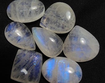 May include: Seven polished moonstone cabochons in various shapes, including oval and teardrop. The stones are translucent with a milky white base and exhibit blue adularescence. The gemstones are displayed against a dark background.