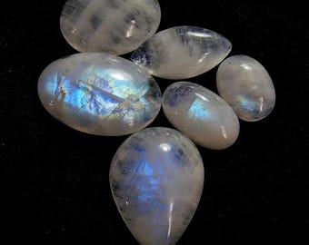 May include: A collection of polished moonstone gemstones in various oval and teardrop shapes. The stones display a milky white base with iridescent blue and rainbow flashes. The gemstones are arranged against a black background.