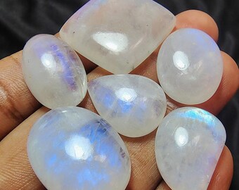 May include: A collection of six polished moonstone gemstones in various shapes, including oval, teardrop, and square. The stones are translucent with a milky white base and exhibit a blue and iridescent sheen. The gemstones are held in a hand.