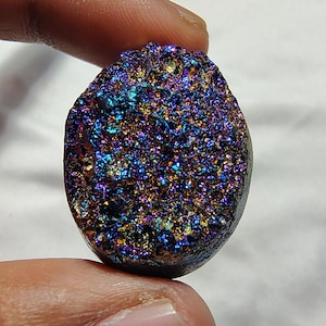 Könnte beinhalten: Ein regenbogenfarbener blauer, violetter und goldener Edelstein mit einer rauen, strukturierten Oberfläche. Der Edelstein ist oval geformt und hat ein natürliches, organisches Aussehen.
