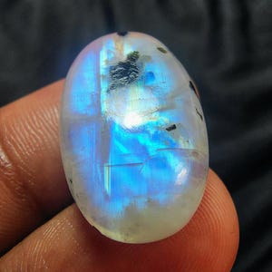 Könnte beinhalten: Ovaler Mondstein-Edelstein mit polierter Oberfläche. Der Stein zeigt ein Farbenspiel mit blauen und weißen Reflexen. Dunkle Einschlüsse sind sichtbar. Der Edelstein wird von einem Finger gehalten.