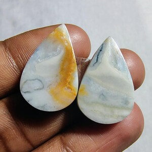 May include: Two teardrop-shaped cabochons of white, blue, and yellow agate. The stones have a swirling pattern and are ready to be set in jewelry.