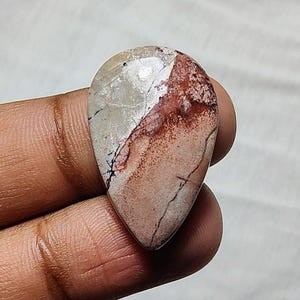 May include: A teardrop-shaped cabochon gemstone with a white, pink, and red pattern. The stone has a smooth, polished surface.