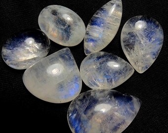 Puede incluir: Siete piedras preciosas de piedra lunar pulidas en varias formas, incluyendo ovaladas y en forma de lágrima. Las piedras tienen una apariencia blanca lechosa translúcida con destellos azules e iridiscentes. Las piedras preciosas se muestran sobre un fondo negro.