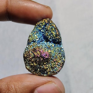 Könnte beinhalten: Ein tropfenförmiger, irisierender Edelstein mit einem Farbverlauf in Blau, Grün, Gold und Lila. Der Edelstein ist glitzernd und hat eine raue, strukturierte Oberfläche.