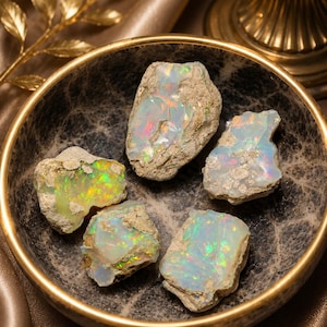 May include: Five raw opal gemstones with iridescent colours are displayed in a decorative marble bowl with a gold rim. The opals have a rough, natural texture and exhibit a range of colours, including green, blue, and red.