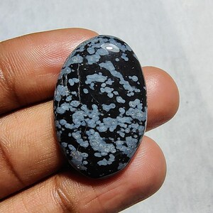May include: A black and white snowflake obsidian cabochon with a smooth, oval shape. The stone has a unique pattern of white snowflake-like inclusions.