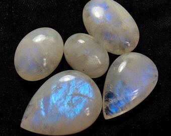 Puede incluir: Cinco piedras de luna pulidas con brillo nacarado e iridiscencia azul. Las piedras varían en tamaño y forma, con un aspecto liso y redondeado. Sobre un fondo negro.