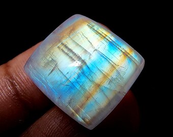 May include: A square-shaped, iridescent moonstone cabochon with a blue and gold sheen. The stone is translucent and has a smooth, polished surface.