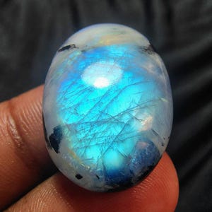 Könnte beinhalten: Ein oval geschliffener Edelstein mit faszinierendem Farbspiel. Der Stein hat hauptsächlich eine blaue Farbe mit schillernden weißen und gelben Reflexen. Interne Brüche sind sichtbar. Der Edelstein wird von einer Person gehalten.