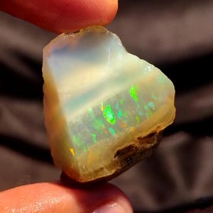 May include: A rough, uncut opal gemstone with a milky white base color. The opal displays flashes of green and yellow iridescence. The stone is held between fingers, showcasing its natural, raw form. The opal's edges are a mix of brown and yellow.