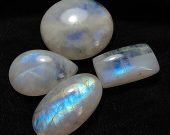 May include: Four polished moonstone gemstones in various shapes: oval, teardrop, and rectangular. The stones display a milky white base with iridescent blue and yellow flashes. The gemstones are set against a black background.