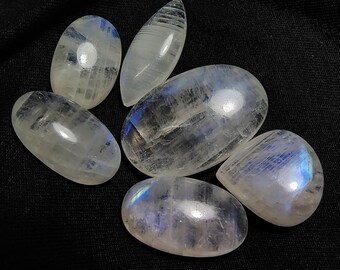 May include: Six polished moonstone gemstones with a translucent, milky white appearance. Each stone has a smooth, rounded shape, with subtle blue and iridescent flashes. The stones are arranged on a black surface.