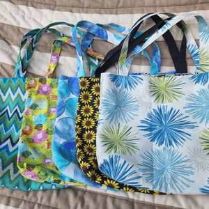 Puede incluir: Cinco bolsas de tela reutilizables con diferentes patrones coloridos. Las bolsas son de diseño sencillo. Los patrones incluyen un chevron azul y verde, un patrón floral verde y rosa, un patrón de acuarela azul, un patrón de girasol negro y amarillo y un fondo blanco con patrones de estallido de estrellas azules y verdes.