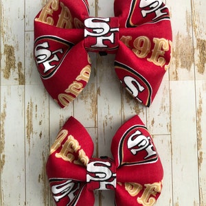 May include: Two red and gold fabric bow ties with the San Francisco 49ers logo. The larger bow tie is on top of the smaller bow tie.