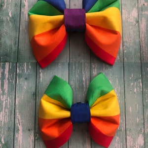 May include: Two rainbow-colored fabric bow ties. The top bow tie has a purple center and the bottom bow tie has a blue center.
