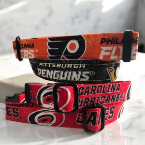 May include: Three dog collars stacked. The top collar is orange with "PHILADELPHIA FLYERS" text and a logo. The middle collar is black with "PITTSBURGH PENGUINS" text. The bottom collar is red with "CAROLINA HURRICANES" text.