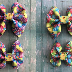 May include: Four Easter-themed hair bows with a colourful pattern of eggs and chicks. The bows are made of fabric and are tied in a bow shape.