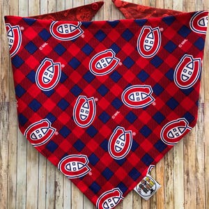 May include: Red and blue plaid dog bandana featuring the Montreal Canadiens logo. The bandana has a red and blue checkered pattern with the team's logo repeated throughout. The bandana is made of fabric and has a tag attached.