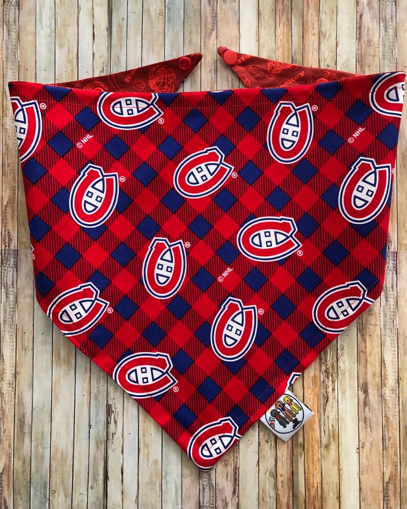 May include: Red and blue plaid dog bandana featuring the Montreal Canadiens logo. The bandana has a red and blue checkered pattern with the team's logo repeated throughout. The bandana is made of fabric and has a tag attached.