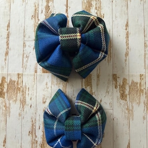 May include: Two blue and green plaid fabric hair bows. The bows are arranged on a light wood grain background. The bows have a central knot and two loops on either side. The plaid pattern includes white, green, and red accents.