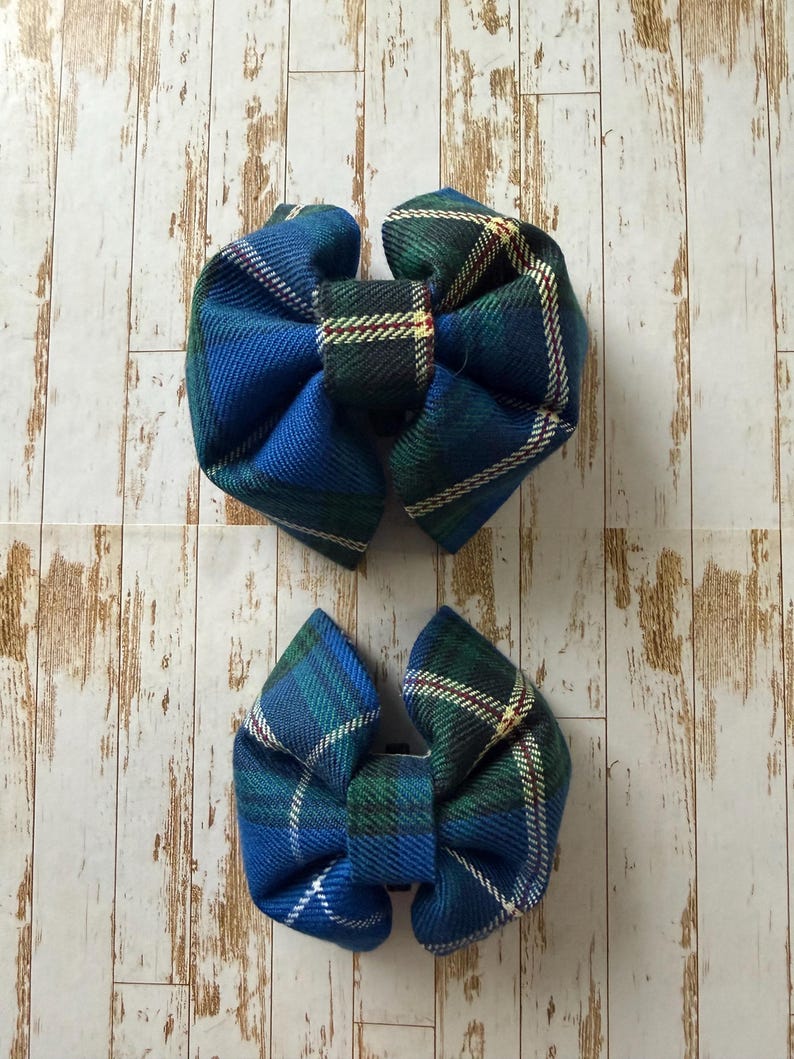May include: Two blue and green plaid fabric hair bows. The bows are arranged on a light wood grain background. The bows have a central knot and two loops on either side. The plaid pattern includes white, green, and red accents.