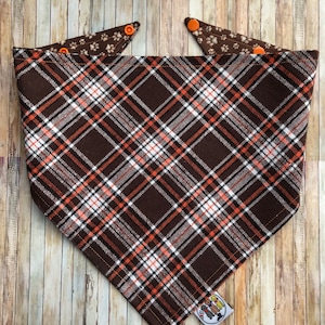 Peut inclure: Un bandana triangulaire pour animal de compagnie avec un motif à carreaux marron, blanc et orange sur le devant. Le revers est marron avec un motif d'empreintes de pattes blanches. Le bandana comprend des boutons-pression en plastique orange.