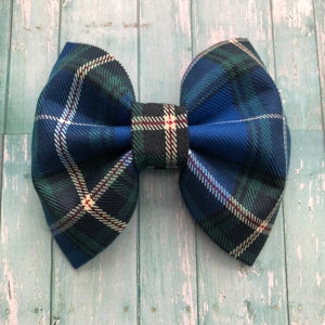 May include: A large, fabric bow in a classic plaid pattern. The bow features a combination of blue, green, and white lines, with a touch of red. The bow is centered with a matching fabric piece. The bow is a hair accessory.