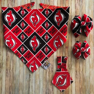 May include: A selection of New Jersey Devils themed pet accessories. Includes a red, black, and white bandana, two bow ties, and a small tie. The items feature the team logo and name on a patterned background.