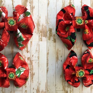 May include: Two sets of red poppy fabric bow ties. The larger bow ties have a white background with red poppies and green leaves. The smaller bow ties have a black background with red poppies and green leaves.
