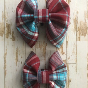 May include: Two plaid bow ties in burgundy, white, and blue. The larger bow tie is on top of the smaller bow tie.