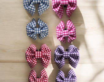 May include: Eight fabric hair bows in a variety of colors. The bows are arranged in two columns of four, with each bow featuring a gingham pattern. Colors include blue, pink, red, and purple. The bows are displayed on a light wooden surface.