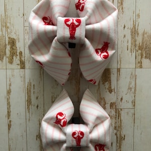 May include: Two white and pink striped fabric bow ties with red lobster illustrations. The larger bow tie is on top of the smaller bow tie.