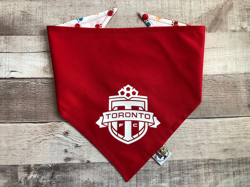 May include: Red dog bandana featuring a white Toronto FC logo. The bandana has a white reverse side with colourful paw prints.