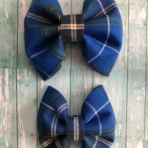 May include: Two blue plaid fabric hair bows. The bows have a classic bow shape with a central knot and rounded loops. The plaid pattern includes shades of blue, green, white, and red. The bows are displayed on a light blue wooden surface.