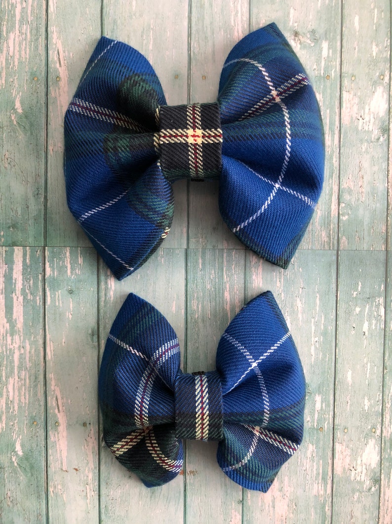 May include: Two blue plaid fabric hair bows. The bows have a classic bow shape with a central knot and rounded loops. The plaid pattern includes shades of blue, green, white, and red. The bows are displayed on a light blue wooden surface.