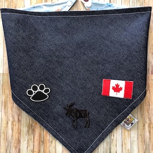 May include: A dark blue denim bandana with a white paw print, a moose silhouette, and a Canadian flag patch. The bandana is folded in a triangle shape and has a white border.