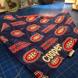 May include: A blue dog bandana featuring the Montreal Canadiens logo in red and white. The bandana has a repeating pattern of the team logo and the words "MONTREAL CANADIENS". A white label with the name "Cooper" is visible.