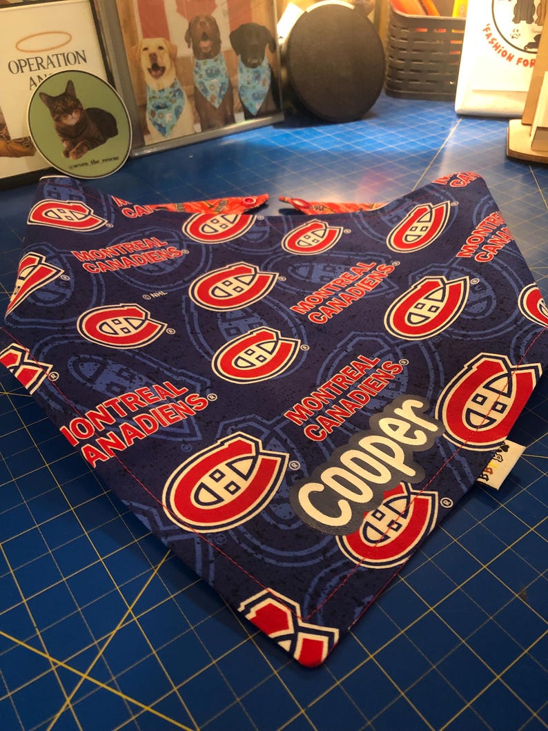 May include: A blue dog bandana featuring the Montreal Canadiens logo in red and white. The bandana has a repeating pattern of the team logo and the words "MONTREAL CANADIENS". A white label with the name "Cooper" is visible.