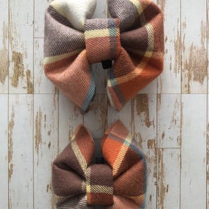 May include: Two plaid fabric hair bows in shades of brown, orange, and blue. The larger bow has a white centre, while the smaller bow has a yellow centre.