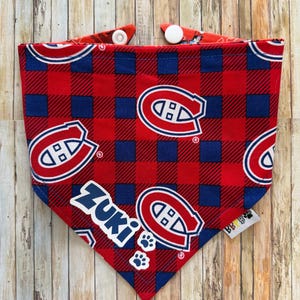May include: Red, blue, and white plaid dog bandana with the text "Zuki" and paw prints. The bandana features the logo of the Montreal Canadiens hockey team. The bandana has white snaps for easy closure.